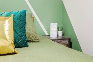 Ein Bett mit einer grünen Bettdecke, einem türkisfarbenen Kissen und einem goldenen Kissen. Auf einem hölzernen Nachttisch steht eine weiße, strukturierte Vase und ein kleines Topf-Pflänzchen.