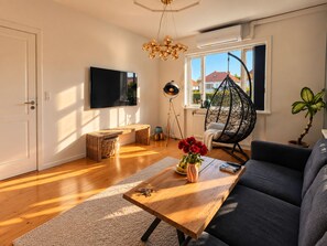 A living room with a wooden coffee table, a sofa, a hanging chair, a television, and a large window with a view of buildings. - Charming Private Apartment in the Heart of København (Copenhagen)