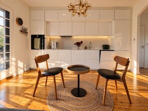 A modern kitchen with white cabinets, a round dining table with two chairs, a chandelier, and a large window. - Charming Private Apartment in the Heart of København (Copenhagen)