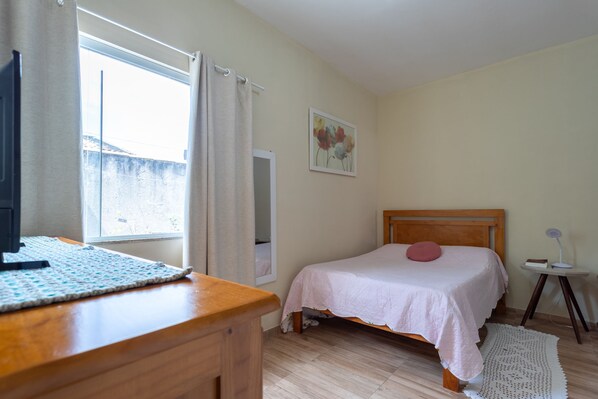 Ein Schlafzimmer mit einem hölzernen Bett, einem Schreibtisch mit Computer, einem kleinen Tisch mit Lampe und einem Fenster mit Vorhängen.