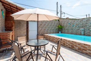 A poolside area with a glass table and chairs, a beige umbrella, and a brick wall. - Private Pool House in California (Cabo Frio)