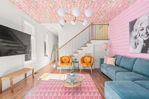 Een woonkamer met een trap, twee oranje fauteuils, een blauwe bank, een houten bankje en een roze vloerkleed met dessin.