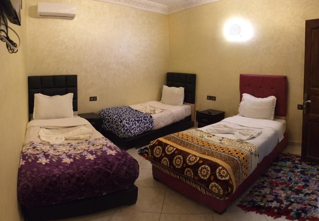 Habitación de hotel con tres camas, cada una con un diseño de cabecera diferente. Se ve un aire acondicionado de pared y una puerta.