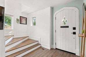 Una escalera conduce a una puerta con un ventanal en forma de rombo, paredes blancas y una pequeña ventana con una planta.