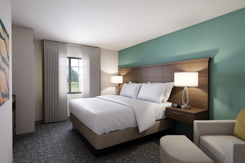 Een hotelkamer met een groot bed, twee bedlampen, een nachtkastje, een stoel en een raam met gordijnen.