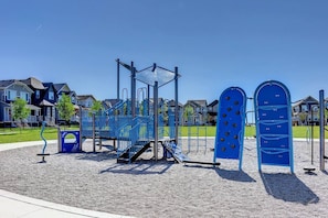 A playground with climbing structures and a slide, surrounded by a gravel area and residential houses in the background. - *2 Bedrooms|Full Kitchen|Faces Park (Calgary)