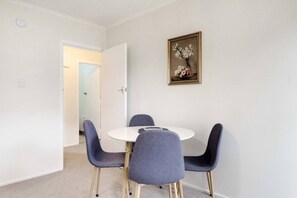 Een kamer met een ronde tafel en vier stoelen, een deur naar een andere kamer, en een lijst met een afbeelding aan de muur.