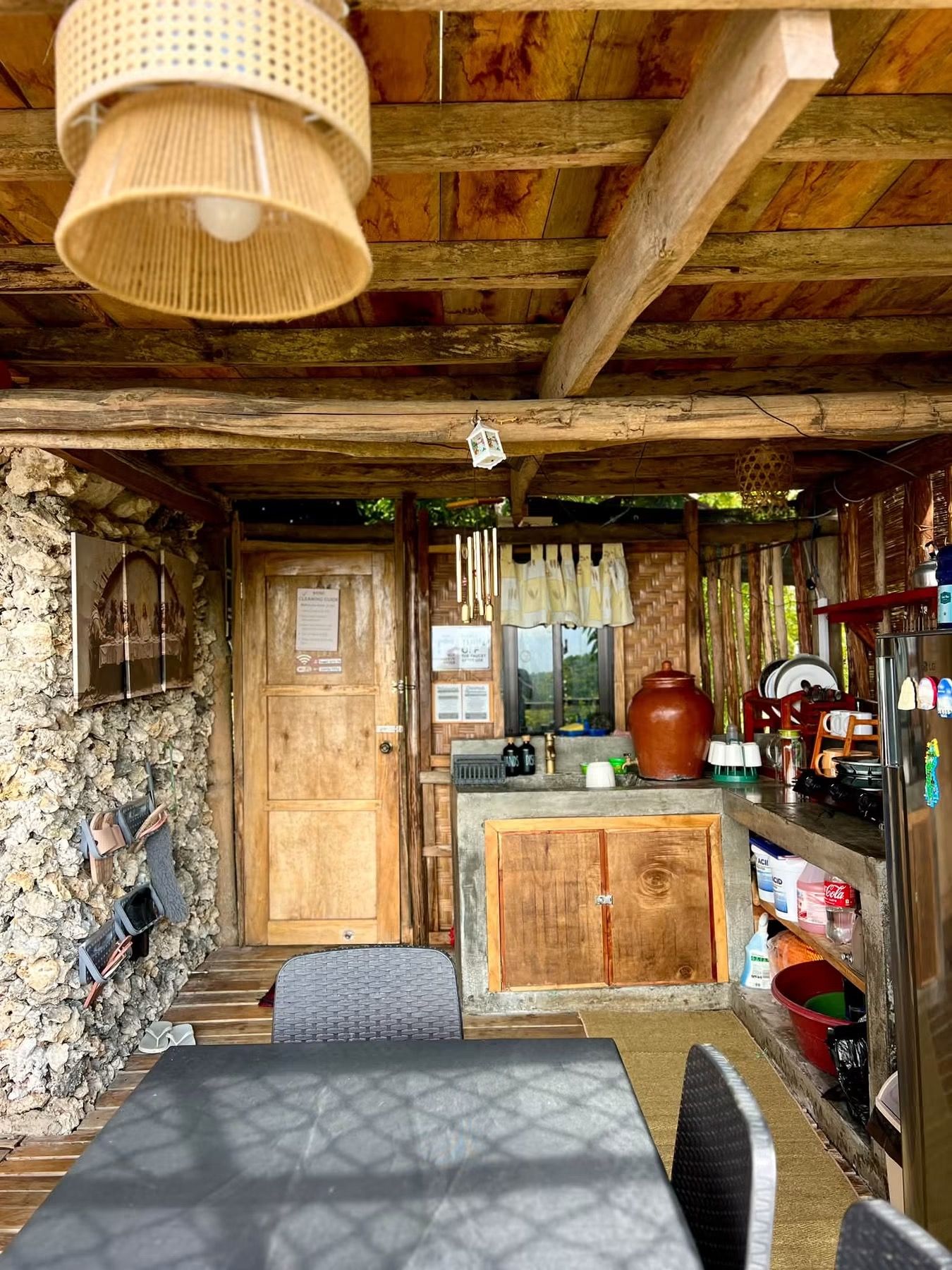 Un intérieur rustique avec des éléments en bois, un mur en pierre, une porte en bois, un coin cuisine équipé d’un réfrigérateur et une table à manger avec des chaises.