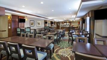 A hotel dining area with multiple tables and chairs, a kitchen in the background, and a flat-screen TV mounted on the wall.