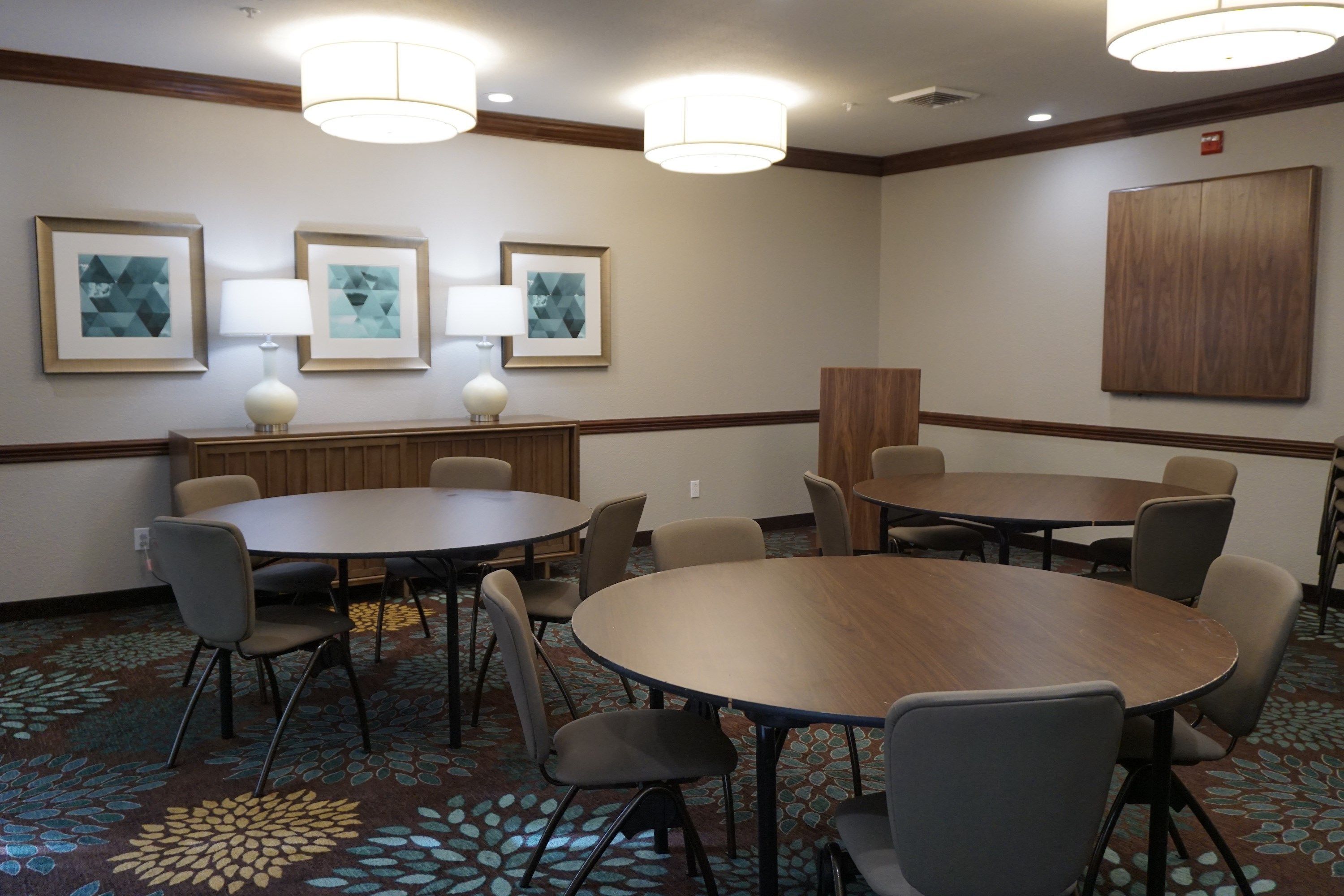 Une salle de conférence avec des tables et des chaises rondes, des œuvres d’art encadrées aux murs et un tapis à motifs.