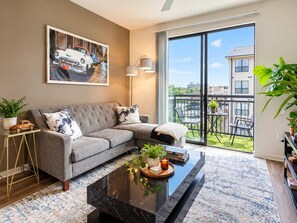 A modern living room with a grey sofa, a coffee table with plants, a painting of a car, and a view of a balcony with outdoor furniture. - The little Havana (Austin)
