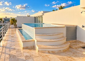 Ein Pool auf dem Dach mit Stufen, umgeben von einer weißen Mauer und wolkenlosem Himmel.