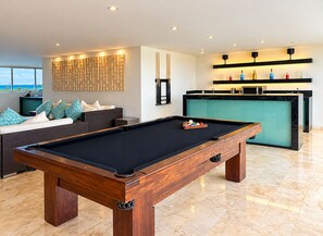 A modern living room with a pool table, a sofa with patterned cushions, a kitchen area with a teal countertop, and a view of the ocean. - Luxury Penthouse-Private Jacuzzi -8-BR (Playa del Carmen)