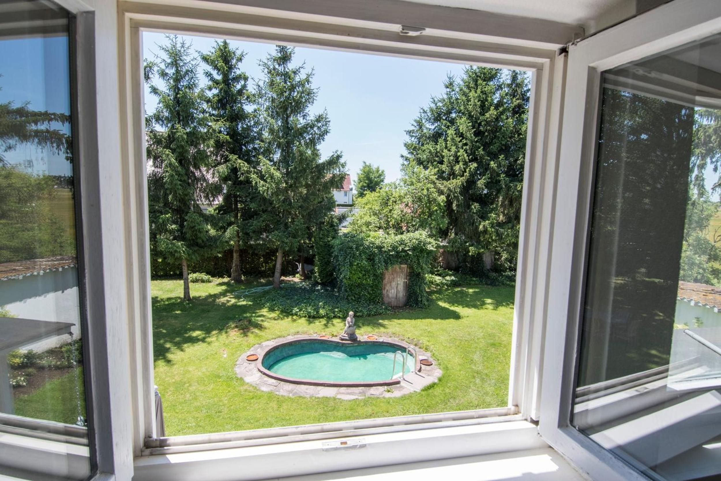 Une vue à travers une fenêtre donnant sur une cour arrière avec une piscine, entourée d’arbres et d’une pelouse verdoyante.