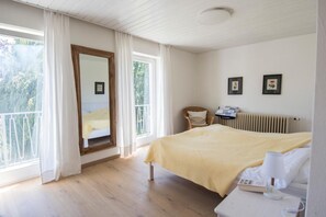 Un dormitorio con cama, una silla, mesita de noche con teléfono y radiador. Hay dos ventanas con cortinas y vistas al exterior con vegetación.