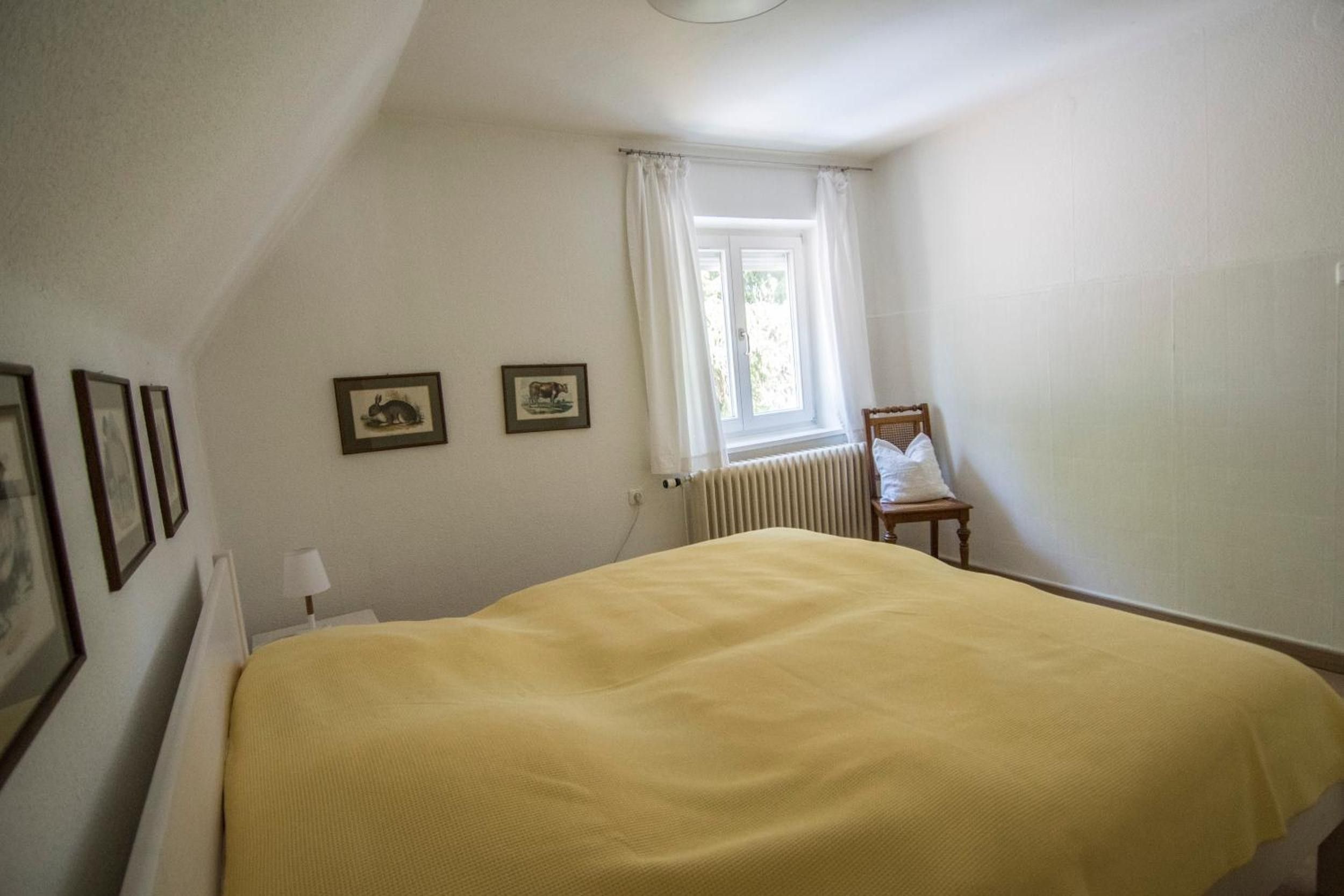 Una camera da letto con un letto, una sedia, una finestra con tende e quadri appesi al muro.