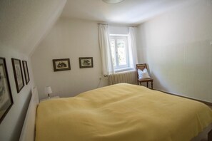 A bedroom with a bed, a chair, a window with curtains, and framed pictures on the wall.