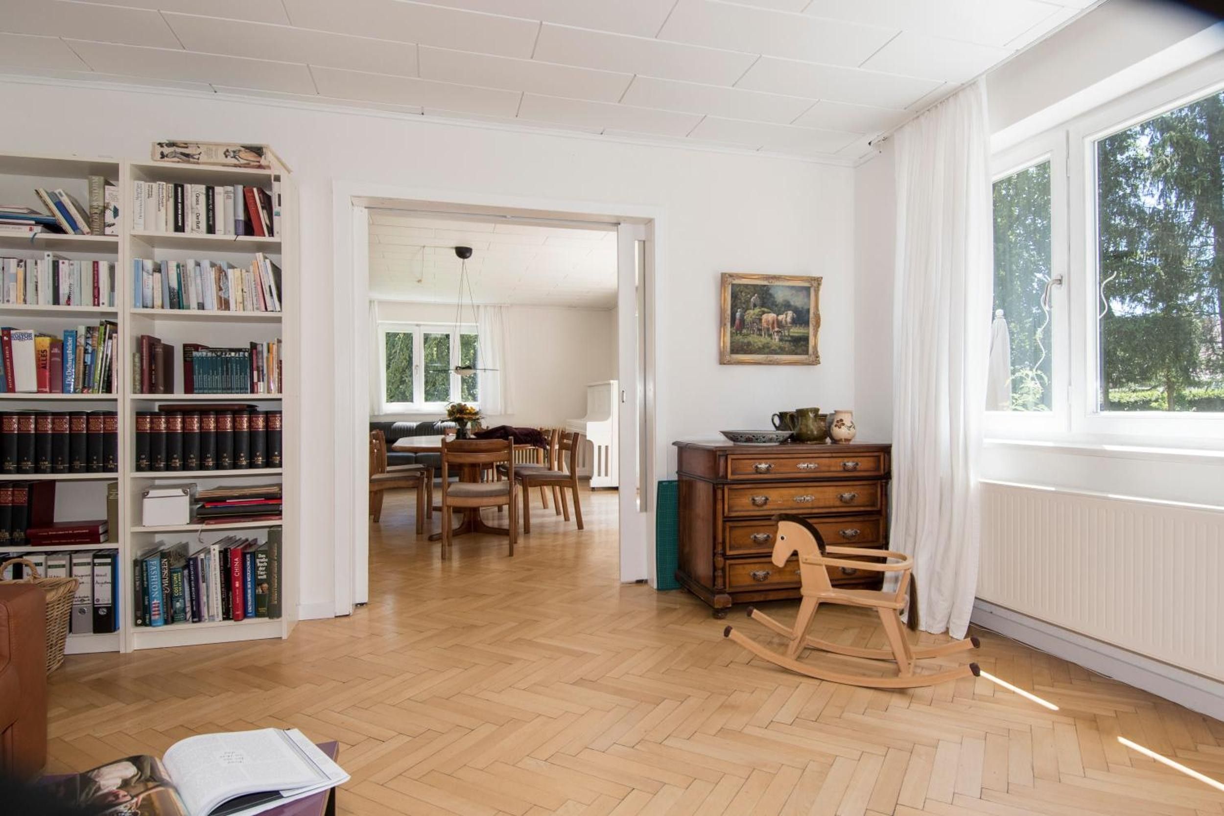 Een ruime kamer met een grote boekenkast, een eethoek en een raam met uitzicht op groen.