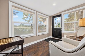 A room with a chess table, two chairs, and large windows with a view of trees and a building. - Modern Waterfront Cottage on Grand River | 4BR (Haldimand)