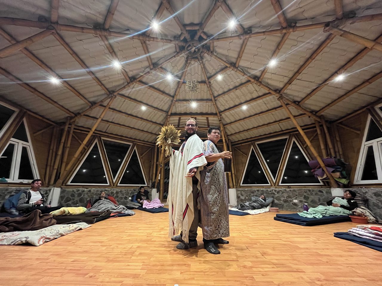 Ein geräumiger, offener Raum mit hohen Decken und Holzbalken, ausgestattet mit Matten und Decken auf dem Boden, und ein paar Personen, die sitzen und liegen.