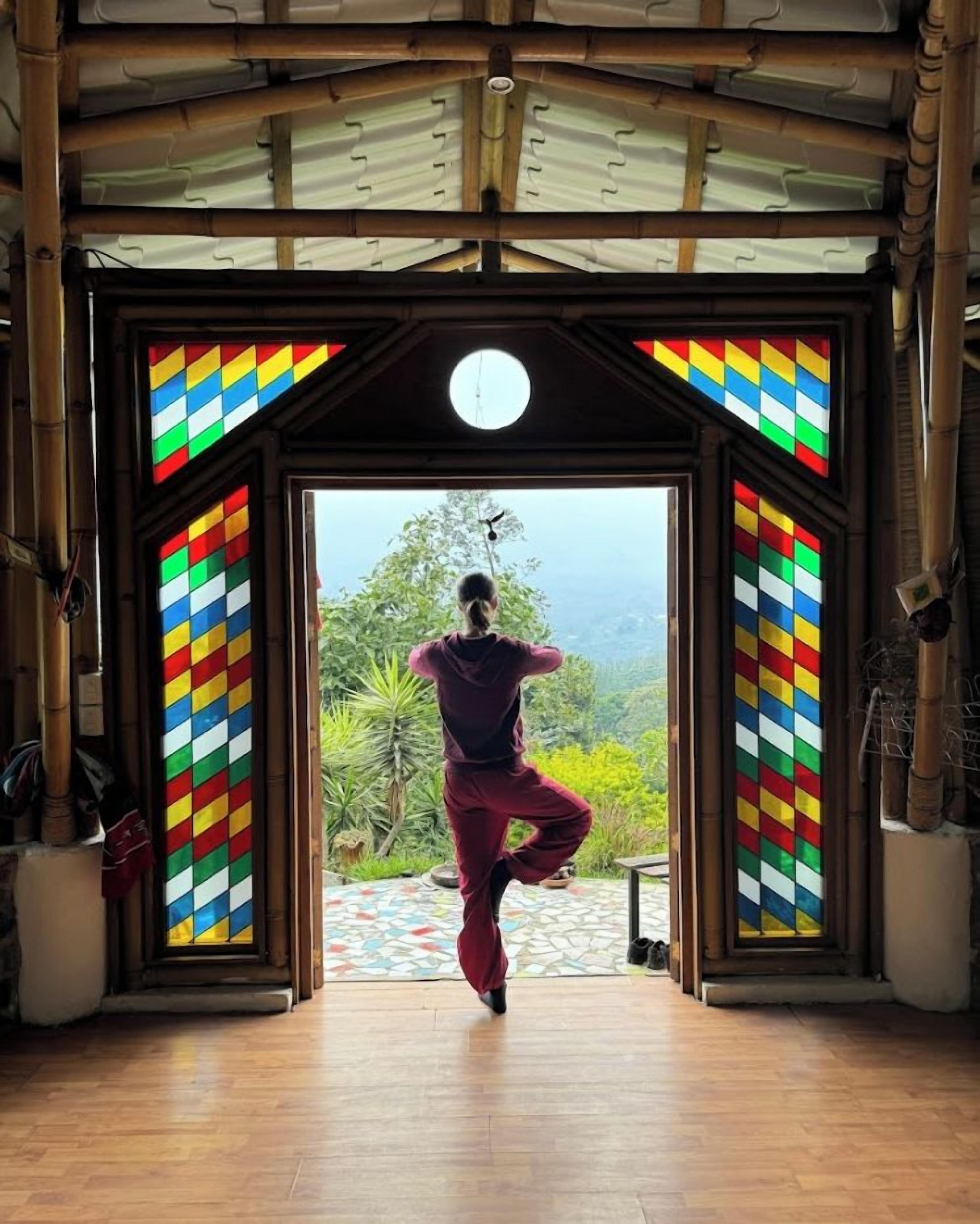 A person performing a yoga pose in a room with a large window, bamboo elements, and a colorful stained glass design.