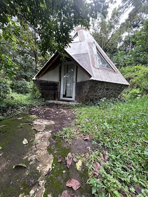 A small stone house with a sloped roof, surrounded by dense greenery and a mossy path leading to the entrance. - Tonkiri Sanctuary (Alotenango)
