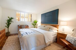 Ein Schlafzimmer mit einem Bett, Nachttischen, einem Schreibtisch, einem Stuhl, einem Fenster mit Jalousien und einem großen Bild an der Wand.