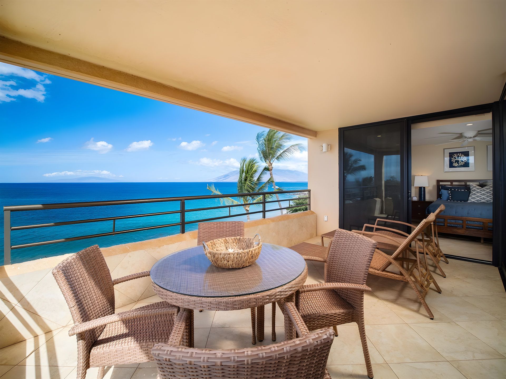 Un balcon avec une table ronde et des chaises en osier, donnant sur une mer d’un bleu limpide et des palmiers.