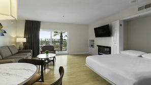 Ein Hotelzimmer mit Bett, Schreibtisch mit Stuhl, Sofa, Fernseher und einem Balkon mit Blick auf Palmen.