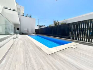 Een modern buitenzwembad met een houten terras, omgeven door een hoge schutting en een gebouw met een glazen gevel.
