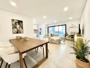 A modern living room with a wooden dining table, white chairs, a sofa, and a large window. - Cabanas de Tavira Strand Apartment (Tavira)