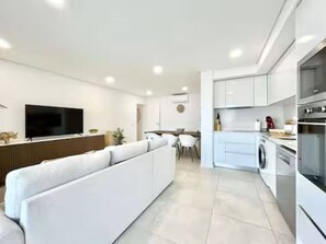 A modern living room with a large white sofa, a flat-screen TV, and a dining area with a table and chairs. - Cabanas de Tavira Strand Apartment (Tavira)
