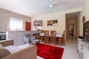A living room with a red rug, a sofa, a dining area with wooden chairs, a kitchen with a stove, and a ceiling fan. - Casa com Piscina Privativa no Calif Rnia (Cabo Frio)