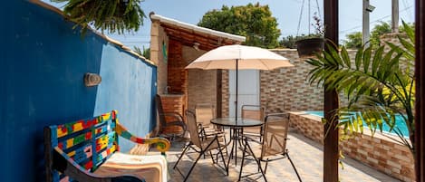 A patio with a blue wall, a colorful bench, a table with chairs, a hanging plant, and a view of a swimming pool.