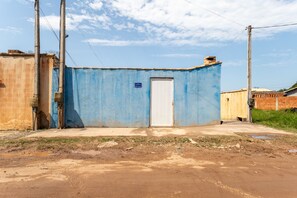 A small, single-story building with a blue exterior and a white door. There are utility poles and wires visible, and the ground appears to be unpaved with some puddles. - Casa com Piscina Privativa no Calif Rnia (Cabo Frio)
