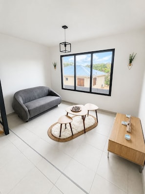 A modern living room with a grey sofa, a small round table with a keyboard, a coffee table, a wooden side table with a candle, and a large window with a view of a house. - The Palmier Bleu Duplex! (Rivière-Salée)