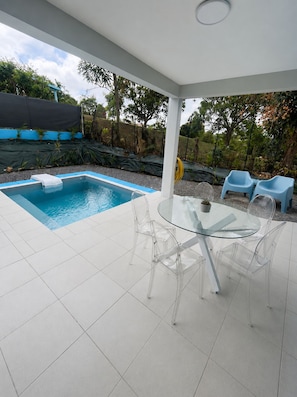 A modern outdoor pool area with a glass table and chairs, surrounded by a white tiled floor and a covered section with blue chairs. - The Palmier Bleu Duplex! (Rivière-Salée)