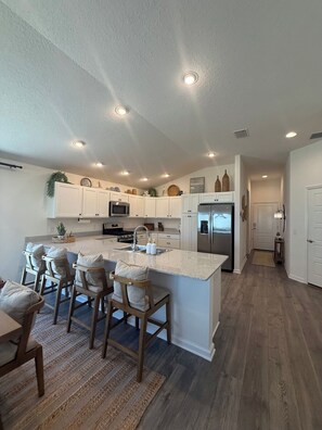 A modern kitchen with white cabinets, a central island with bar stools, and a stainless steel refrigerator. - Brand-New Stylish 2BR Villa with Golf Cart in The Villages (The Villages)