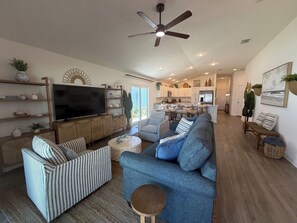 A spacious living room with a large flat-screen TV, a ceiling fan, and a kitchen in the background. - Brand-New Stylish 2BR Villa with Golf Cart in The Villages (The Villages)