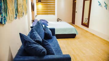 Una stanza con un divano blu, un letto e un pavimento in legno. Si vede una finestra con persiane e una porta con uno specchio.