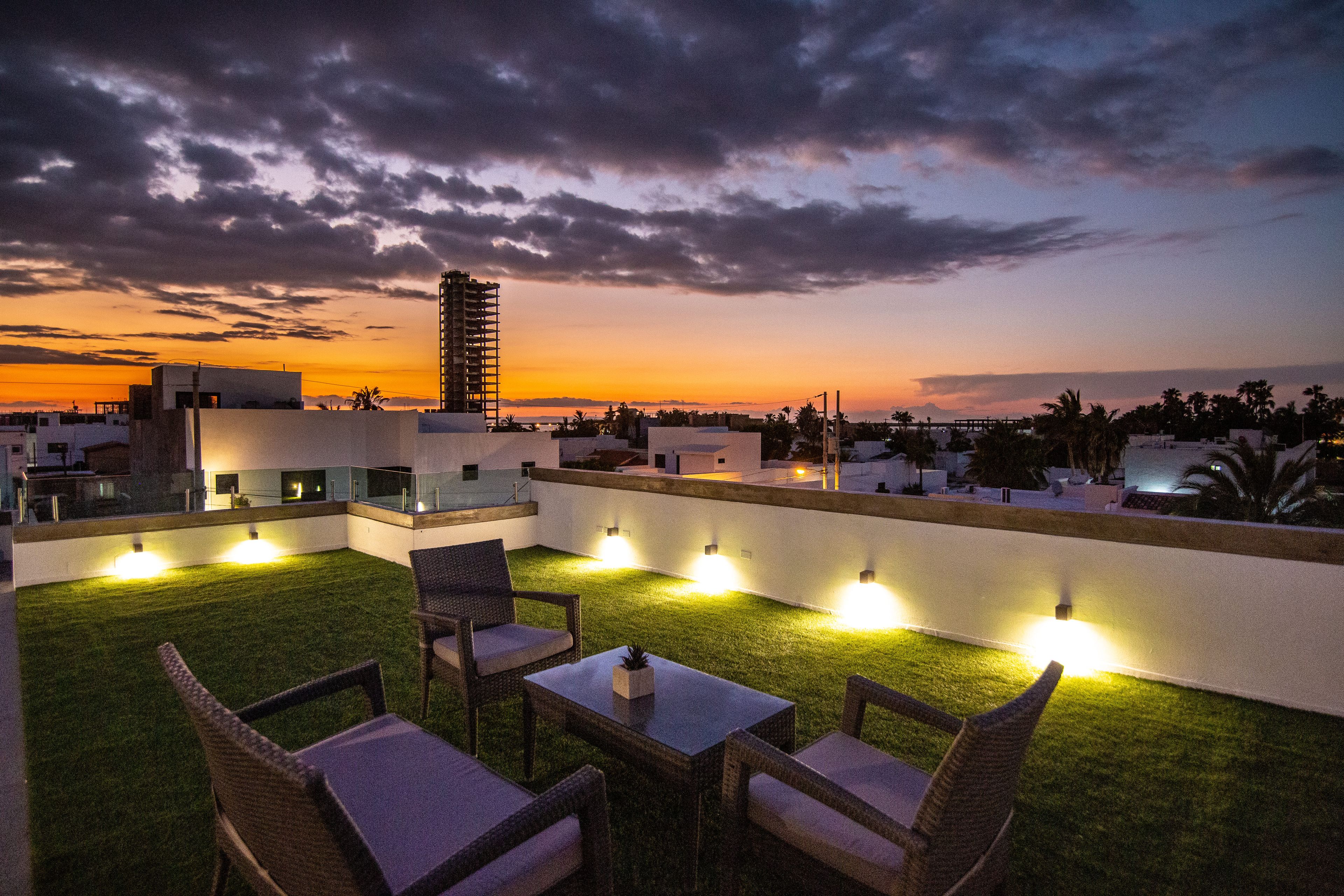 Un área de estar al aire libre con sillas y mesas, un césped y vistas del horizonte de la ciudad al atardecer.