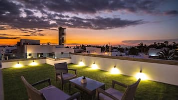 An outdoor seating area with chairs and tables, a grassy lawn, and a view of the city skyline at sunset.