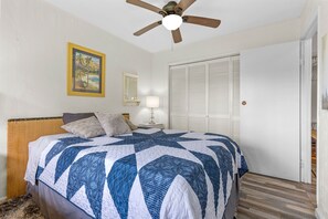 A bedroom with a bed, a ceiling fan, a nightstand with a lamp, a door, and a framed picture on the wall. - Peaceful 2BR Cottage in Keaau | Volcano & Beaches (Keaau)