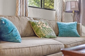 A sofa with three pillows, one with a leaf pattern and text, and two solid color pillows. A lamp is on a side table next to the sofa. - Peaceful 2BR Cottage in Keaau | Volcano & Beaches (Keaau)