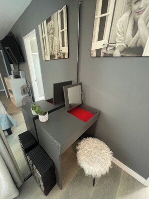 Une pièce moderne avec un bureau gris, un siège rembourré rouge, un tabouret blanc et moelleux, une petite plante et un téléviseur posé sur un meuble.