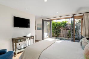 Ein Schlafzimmer mit einem großen Bett, einem an der Wand befestigten Fernseher, einer Holzkommode und einer Glasschiebetür, die auf einen Balkon mit Whirlpool führt.
