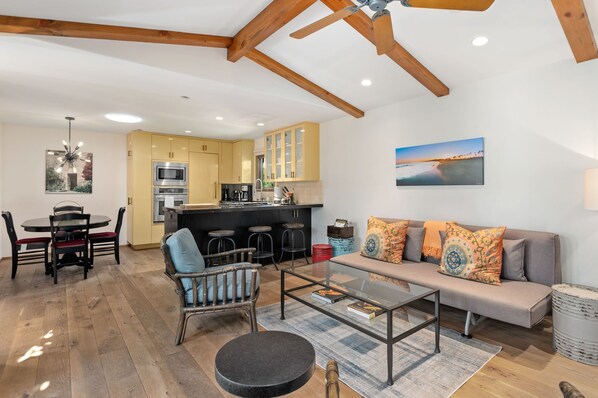 Eine moderne Küche mit integrierten Geräten, ein Essbereich mit rundem Tisch und Stühlen, eine Couch mit gemusterten Kissen und ein Strandbild an der Wand.