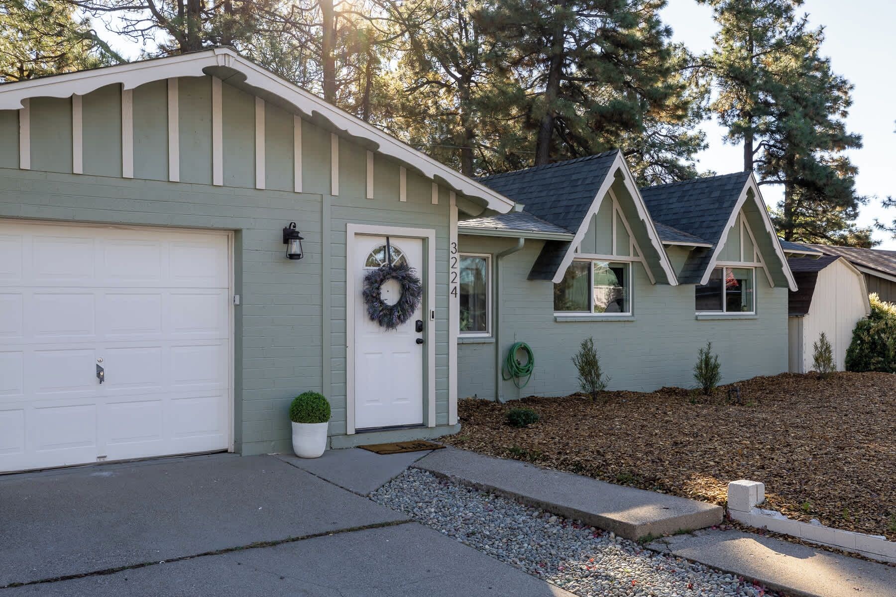 A single-story house with a garage, a front door with a wreath, and a small front yard with mulch and a potted plant.