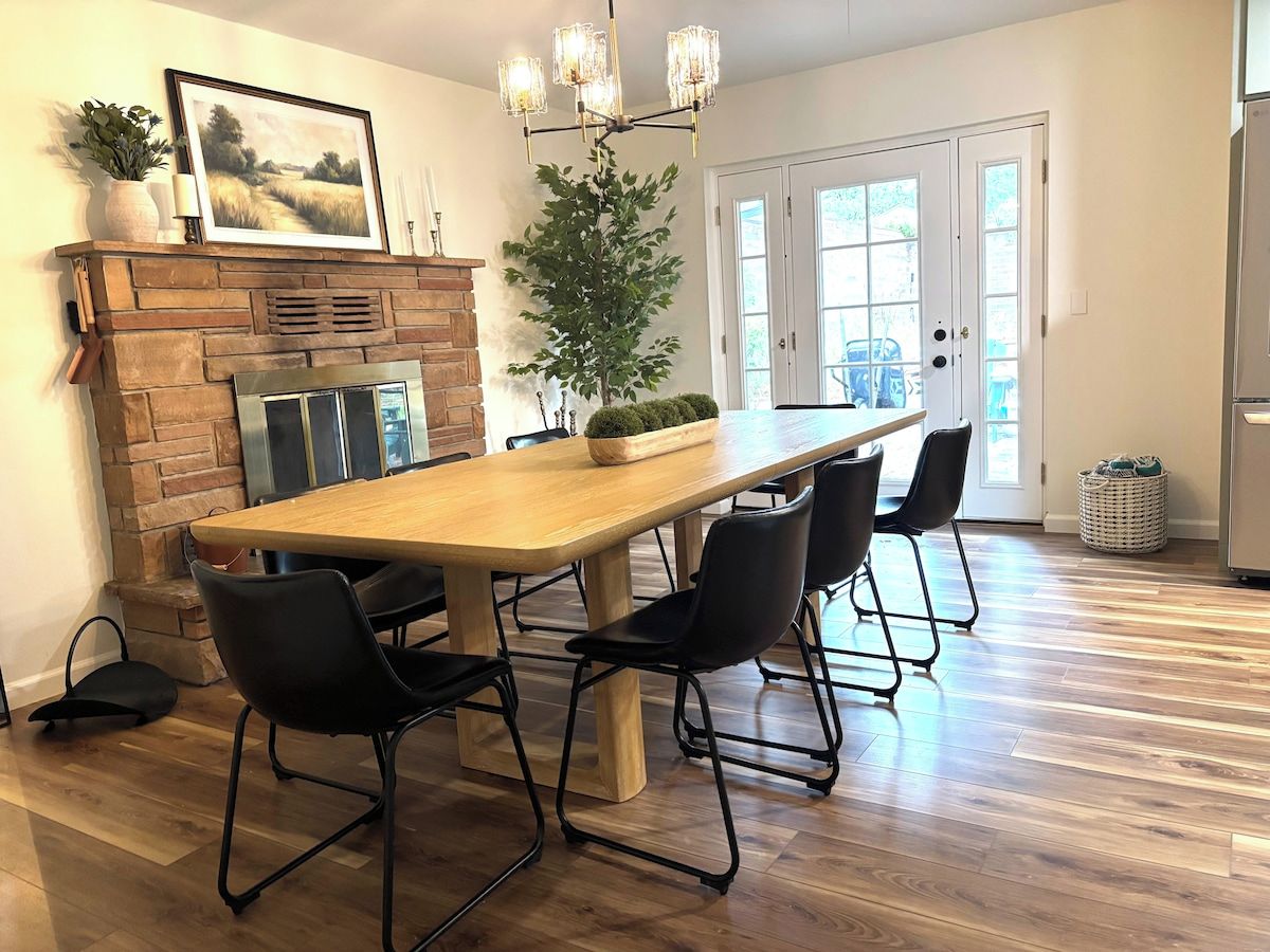 A dining area with a long wooden table, black chairs, a brick fireplace, and a large potted plant.