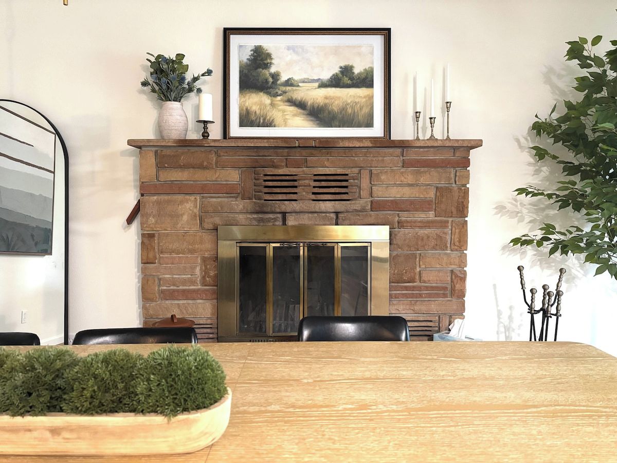 A dining area with a stone fireplace, a framed landscape painting, and a potted plant.